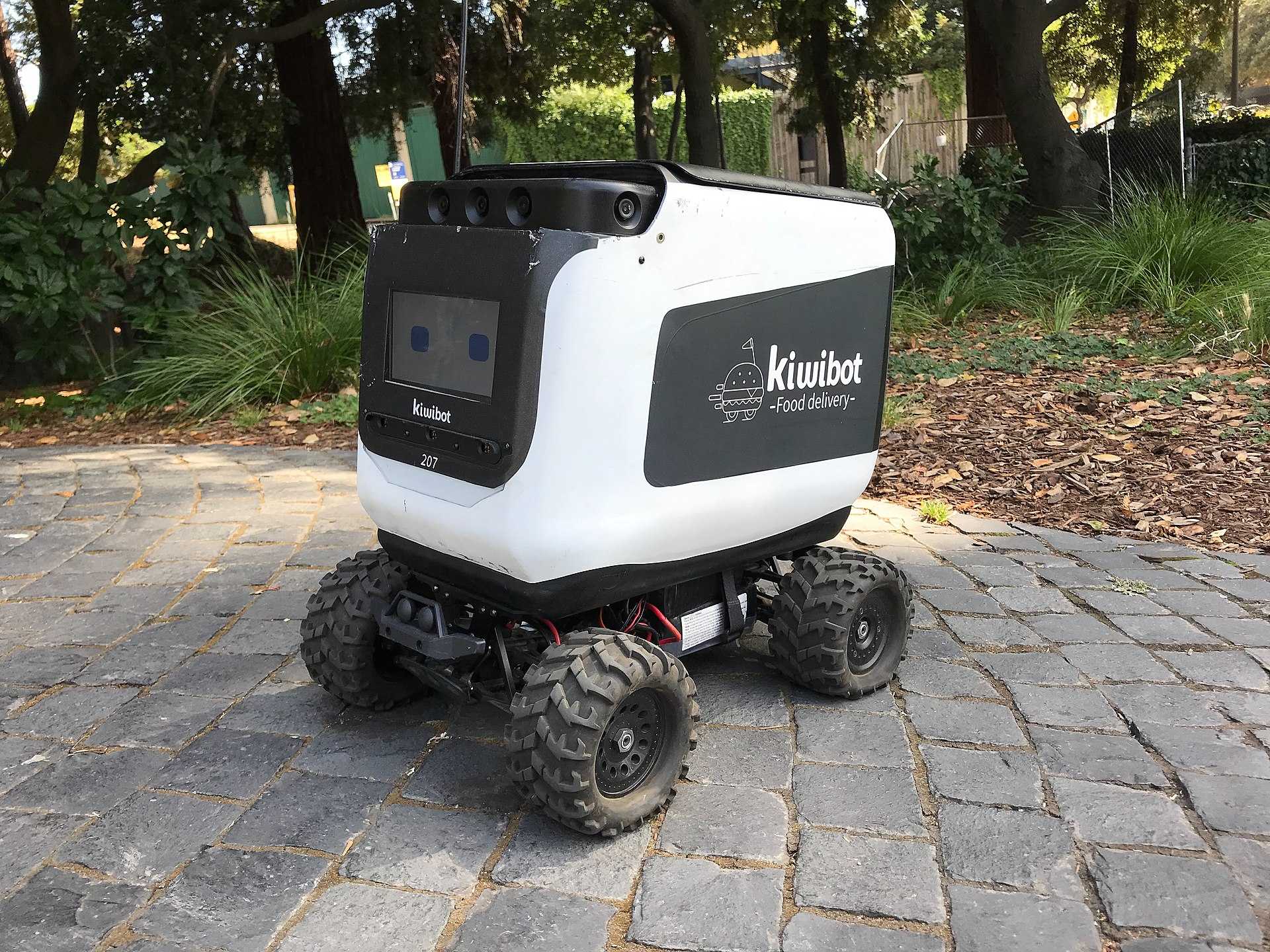 A Kiwibot delivery robot sits on a sidewalk of pavers.