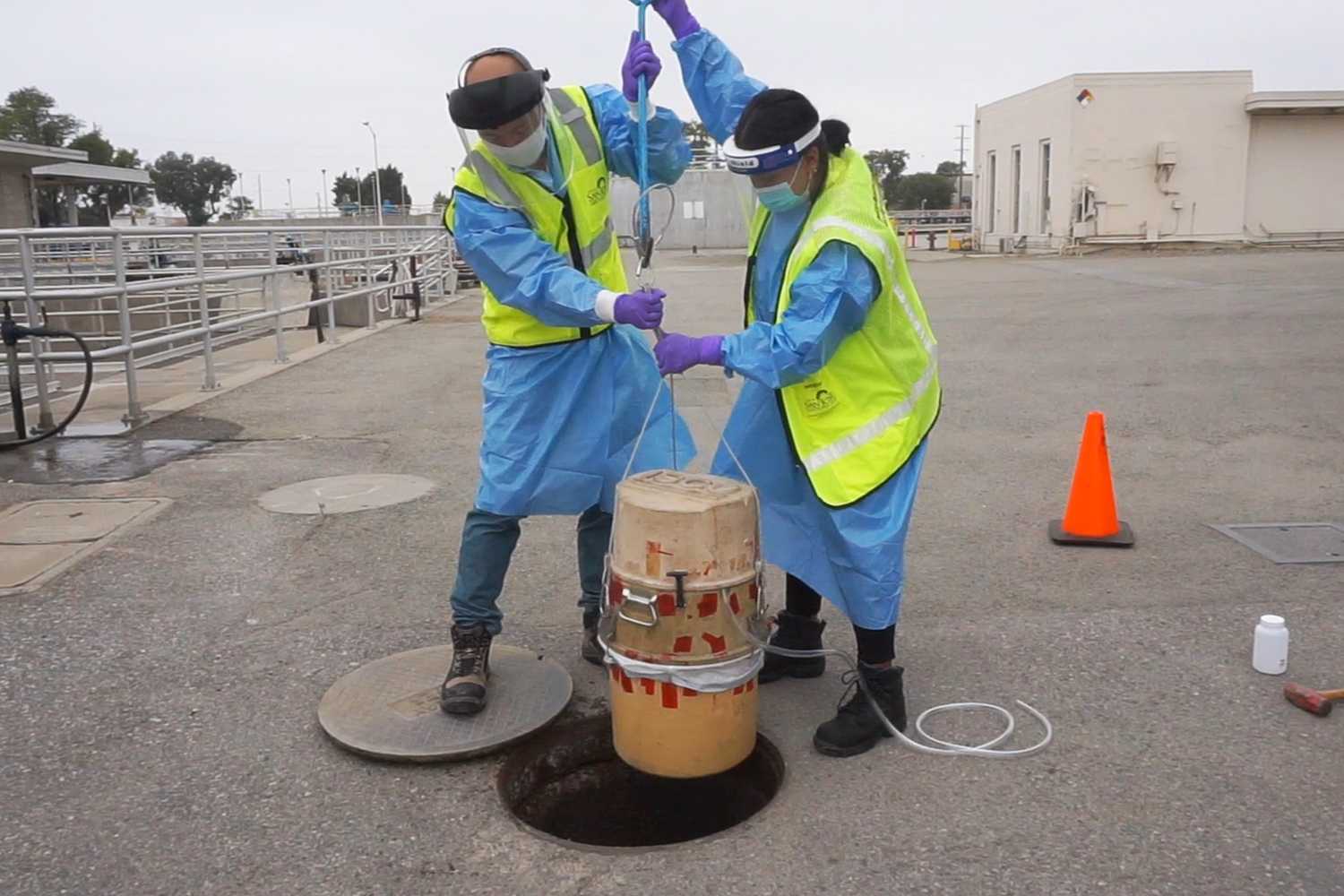 Two technicians hoist a water sample container from an uncovered manhole.