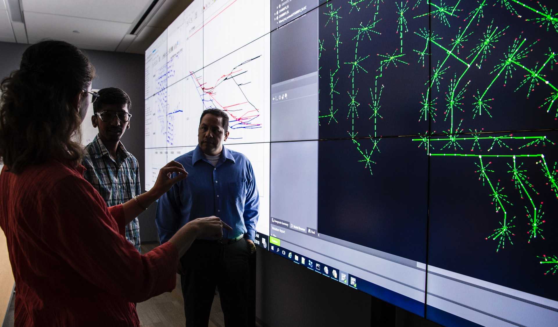 Three researchers discuss a wall-sized display of an energy grid simulation.