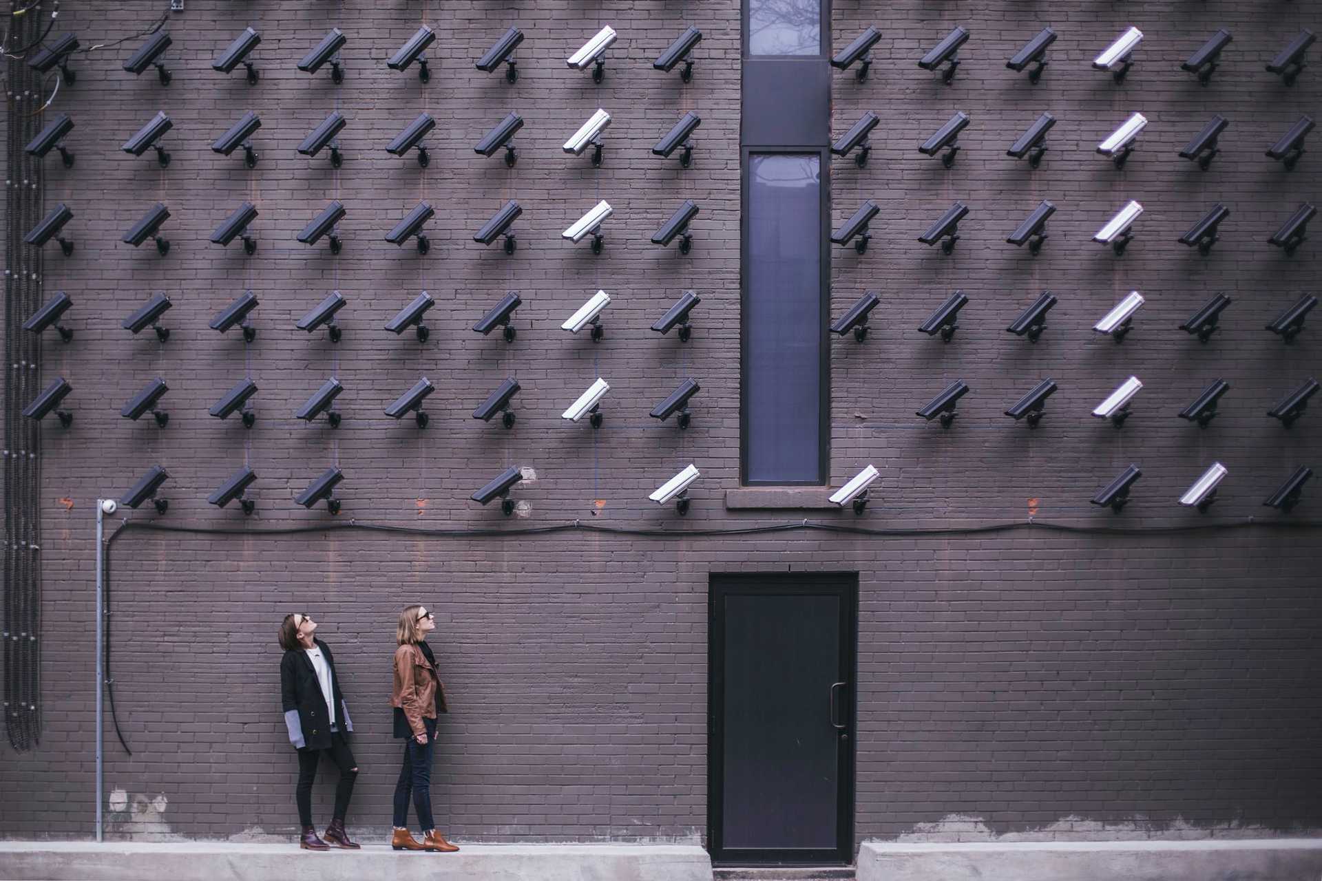 Two people stand in front of a wall full of surveillance cameras.